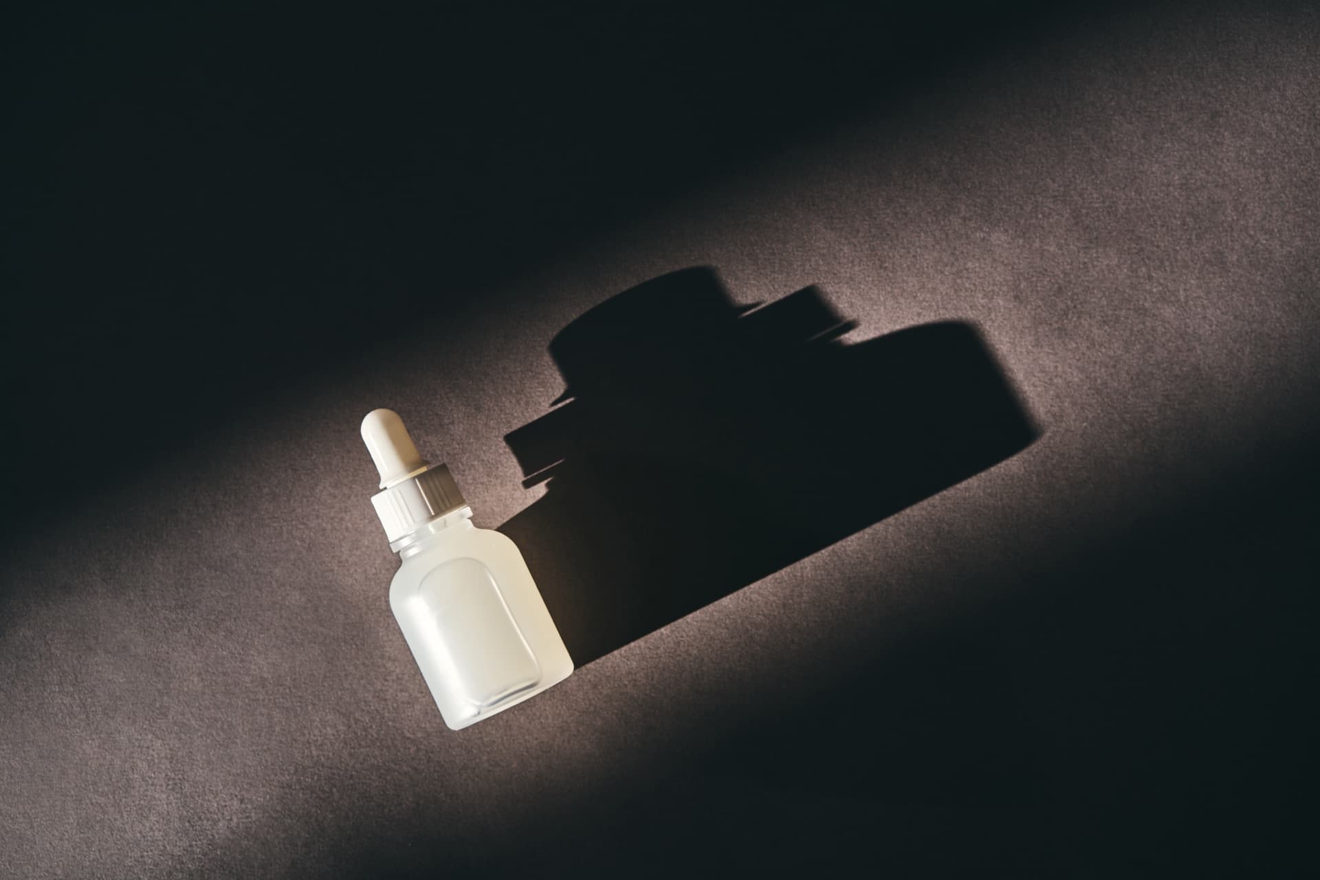 暗色背景中一支白色滴管精華瓶置於一道光束下、投出長長陰影,象徵保養品的保存與光線、溫度的關係